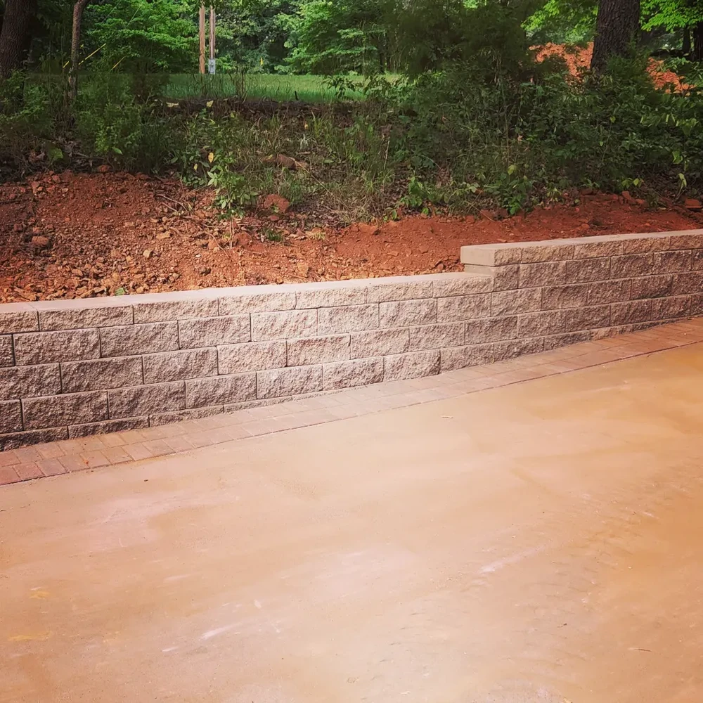 Tan block retaining wall holding back red clay soil at the edge of a wooded backyard.