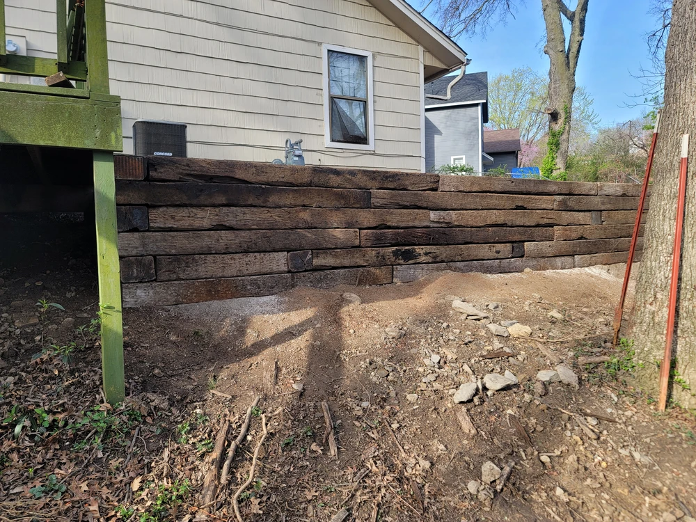 Gray block retaining wall along a metal building with bare soil behind it and a concrete walkway nearby.